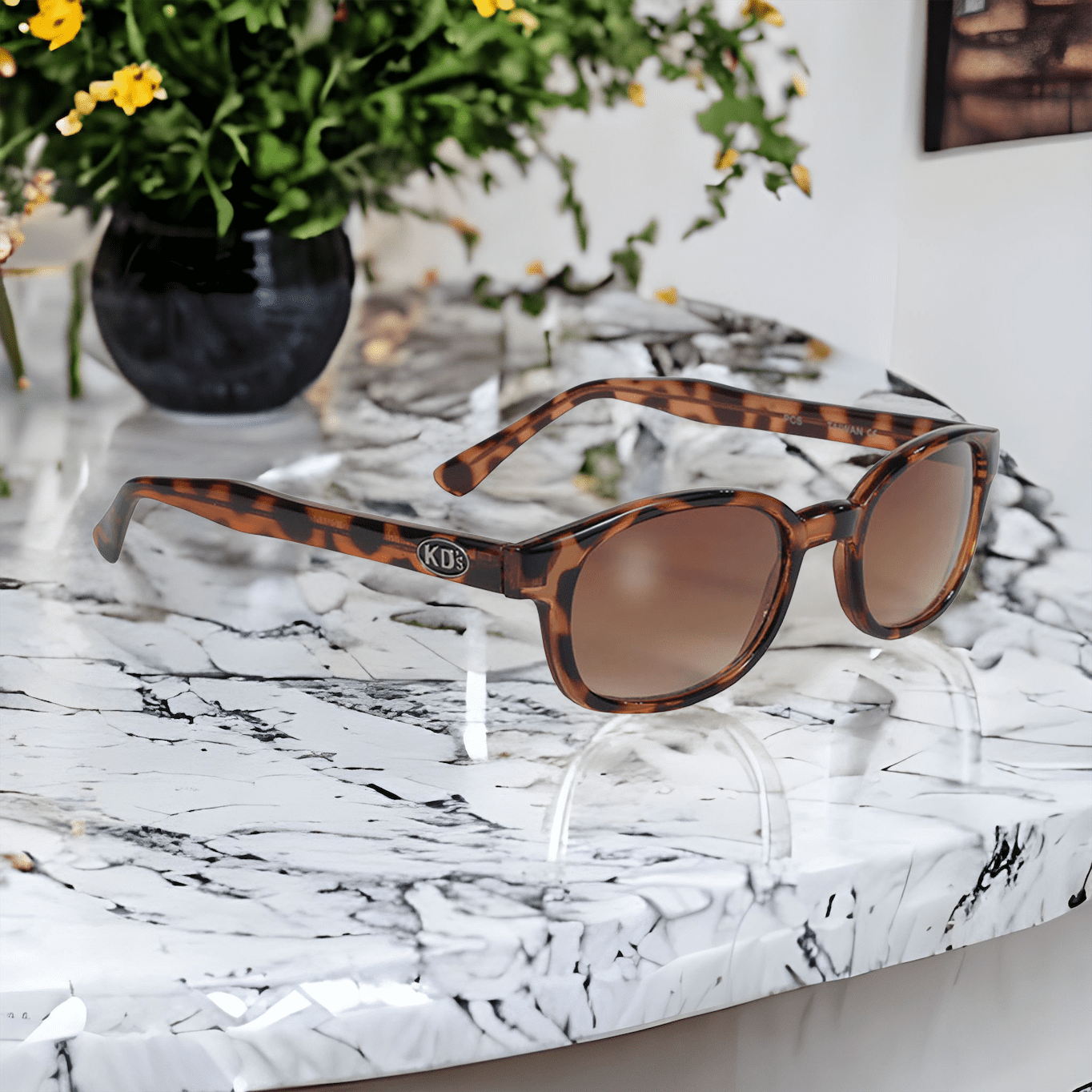 Lunettes de soleil KD's 200 avec des verres ambrés et un cadre décoré façon écaille de tortue convenant aux motards et aux sportifs sur un comptoir en marbre blanc devant une plante.