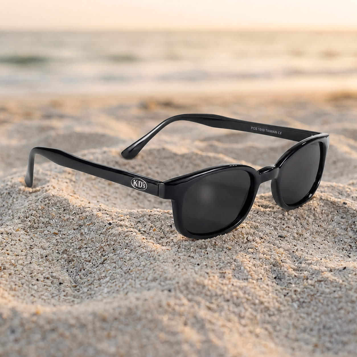 Lunettes de soleil X-KD's 1010 noir brillant à verres fumés, monture grande taille, photographiées sur un sable clair en bord de mer.