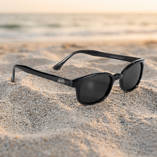 Lunettes de soleil X-KD's 1010 noir brillant à verres fumés, monture grande taille, photographiées sur un sable clair en bord de mer.