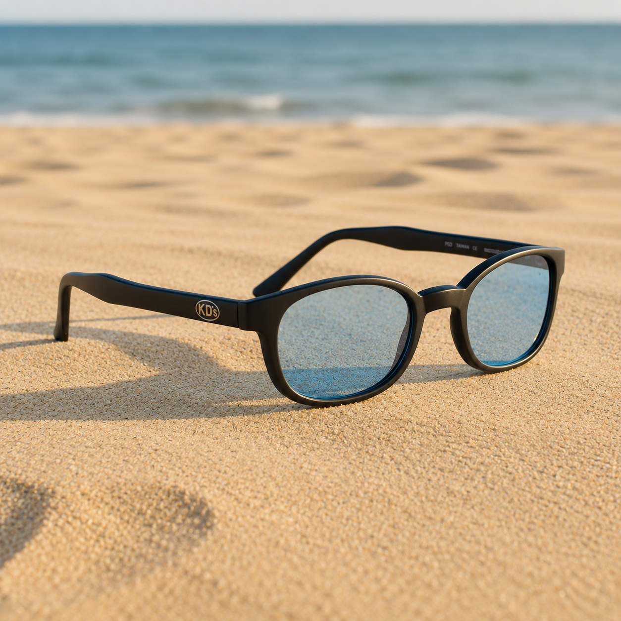 Lunettes de soleil KD's 20012 à monture noir mat et verres bleu ciel posées sur une plage de sable face à la mer.