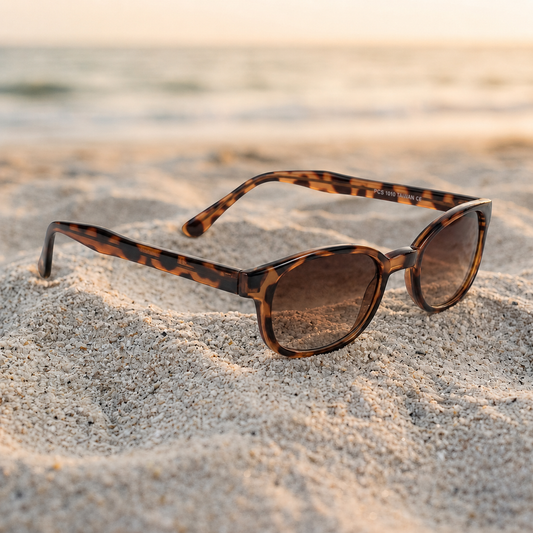Lunettes de soleil KD's 200 à monture écaille brune et verres ambre posées sur une plage de sable face à la mer.