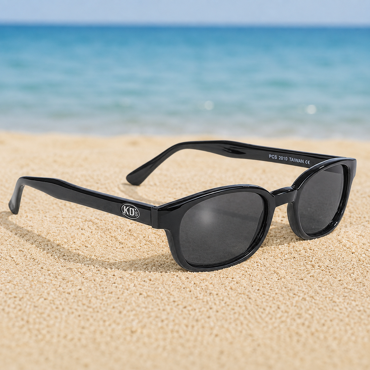 Lunettes de soleil KD's 2010 à monture noir brillant et verres fumés posées sur une plage de sable face à la mer.