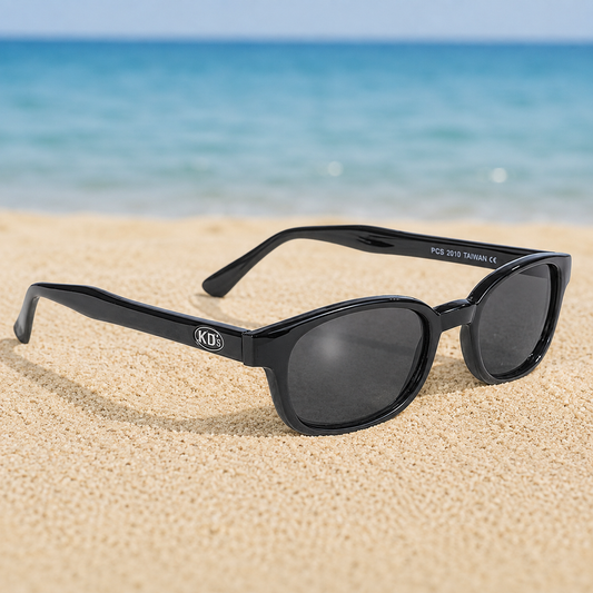 Lunettes de soleil KD's 2010 à monture noir brillant et verres fumés posées sur une plage de sable face à la mer.