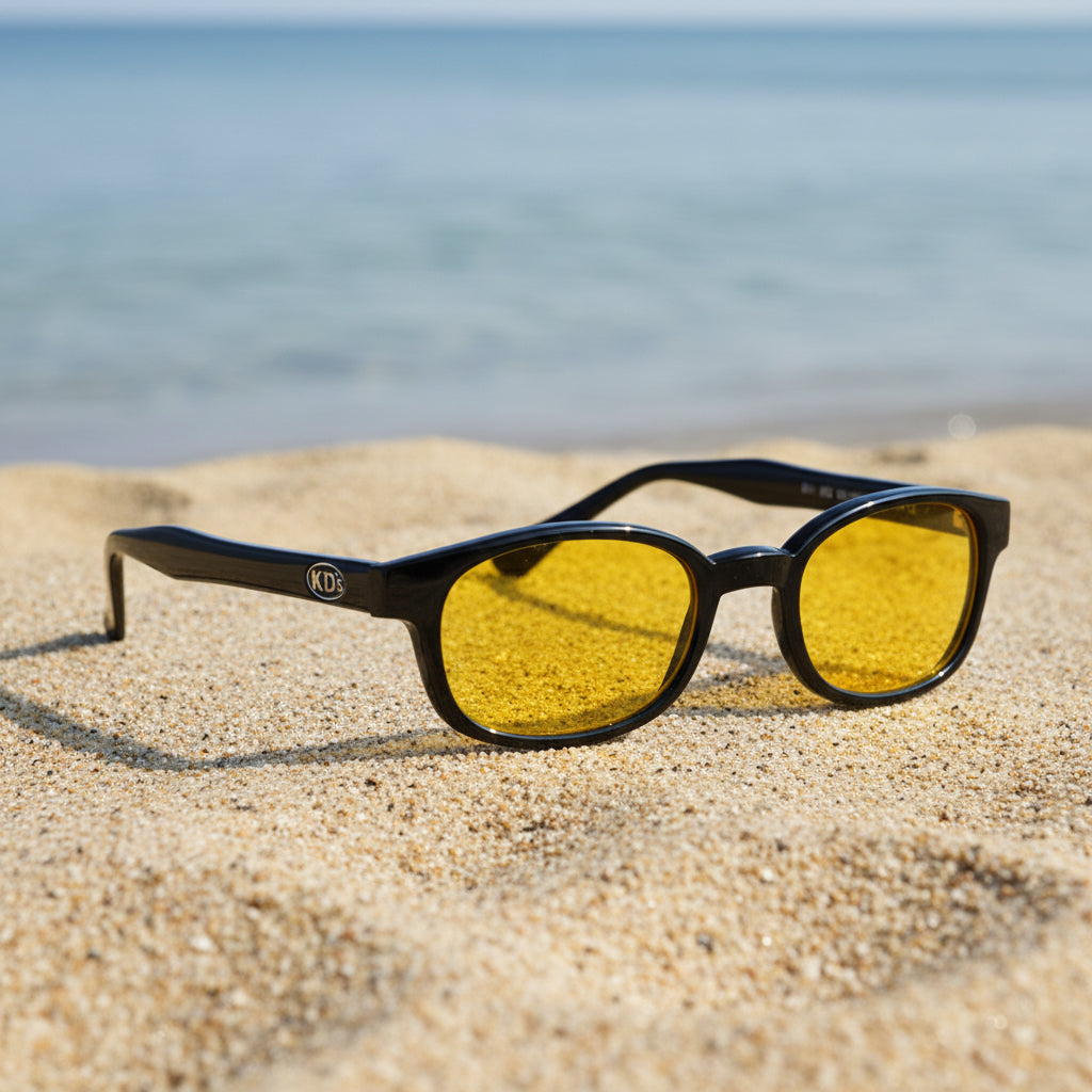 Lunettes KD's 21112 à monture noir brillant et verres jaunes posées sur une plage de sable face à la mer.
