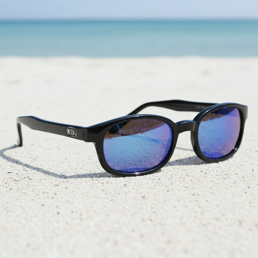Lunettes KD's 20118 à monture noir brillant et verres miroir colorés irisés bleu-violet posées sur une plage de sable face à la mer.