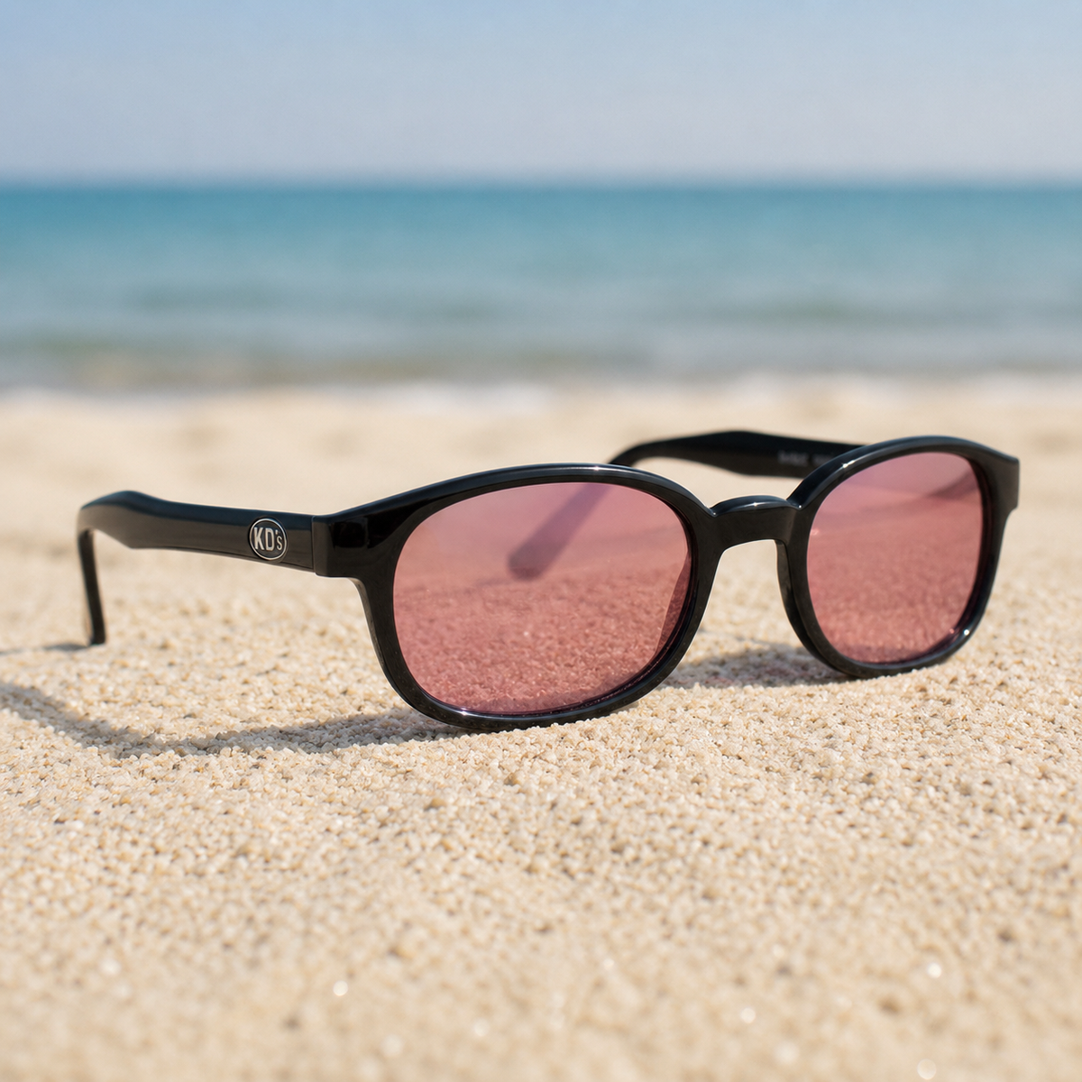 Lunettes KD's 20120 à monture noir brillant et verres rose sombre légèrement transparents posées sur une plage de sable face à la mer.
