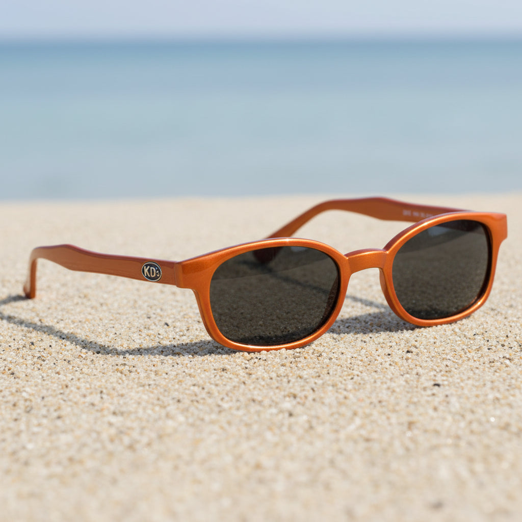 Lunettes de soleil KD's 20128 à monture orange mat et verres fumés posées sur une plage de sable face à la mer.