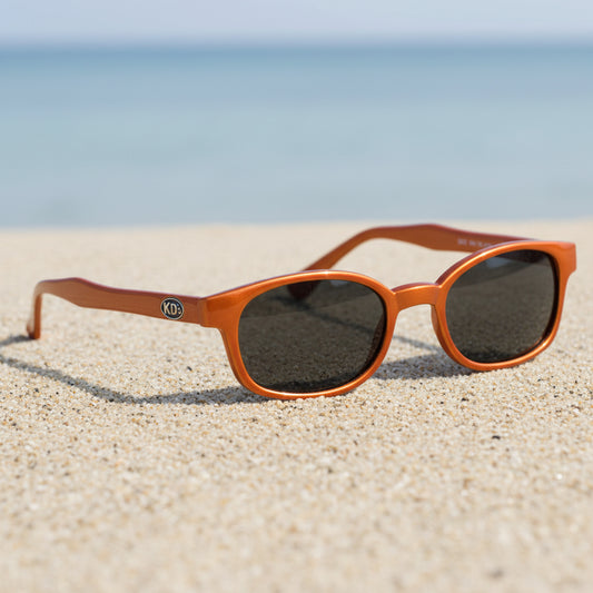 Lunettes de soleil KD's 20128 à monture orange mat et verres fumés posées sur une plage de sable face à la mer.