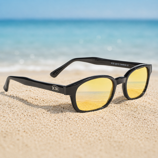 Lunettes KD's 20129 à monture noir brillant et verres jaunes polarisés posées sur une plage de sable face à la mer.