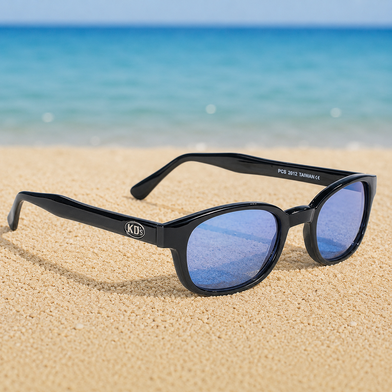 Lunettes de soleil KD's 2012 à monture noir brillant et verres bleus posées sur une plage de sable face à la mer.