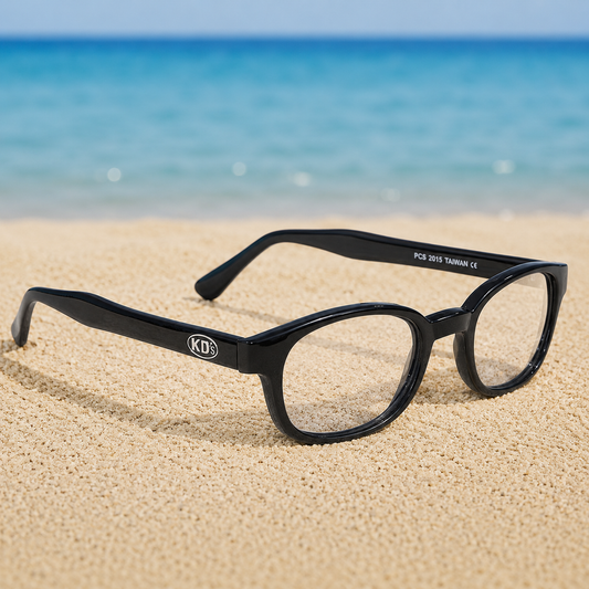 Lunettes de soleil KD's 2015 à monture noir brillant et verres clairs transparents posées sur une plage de sable face à la mer.
