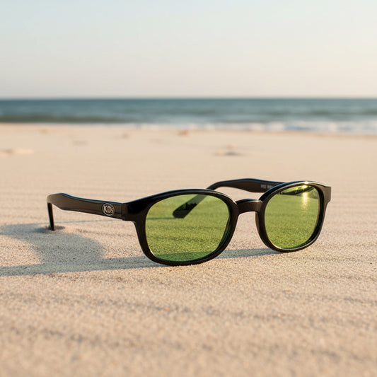 Lunettes de soleil KD's 2016 à monture noir brillant et verres verts posées sur une plage de sable face à la mer.