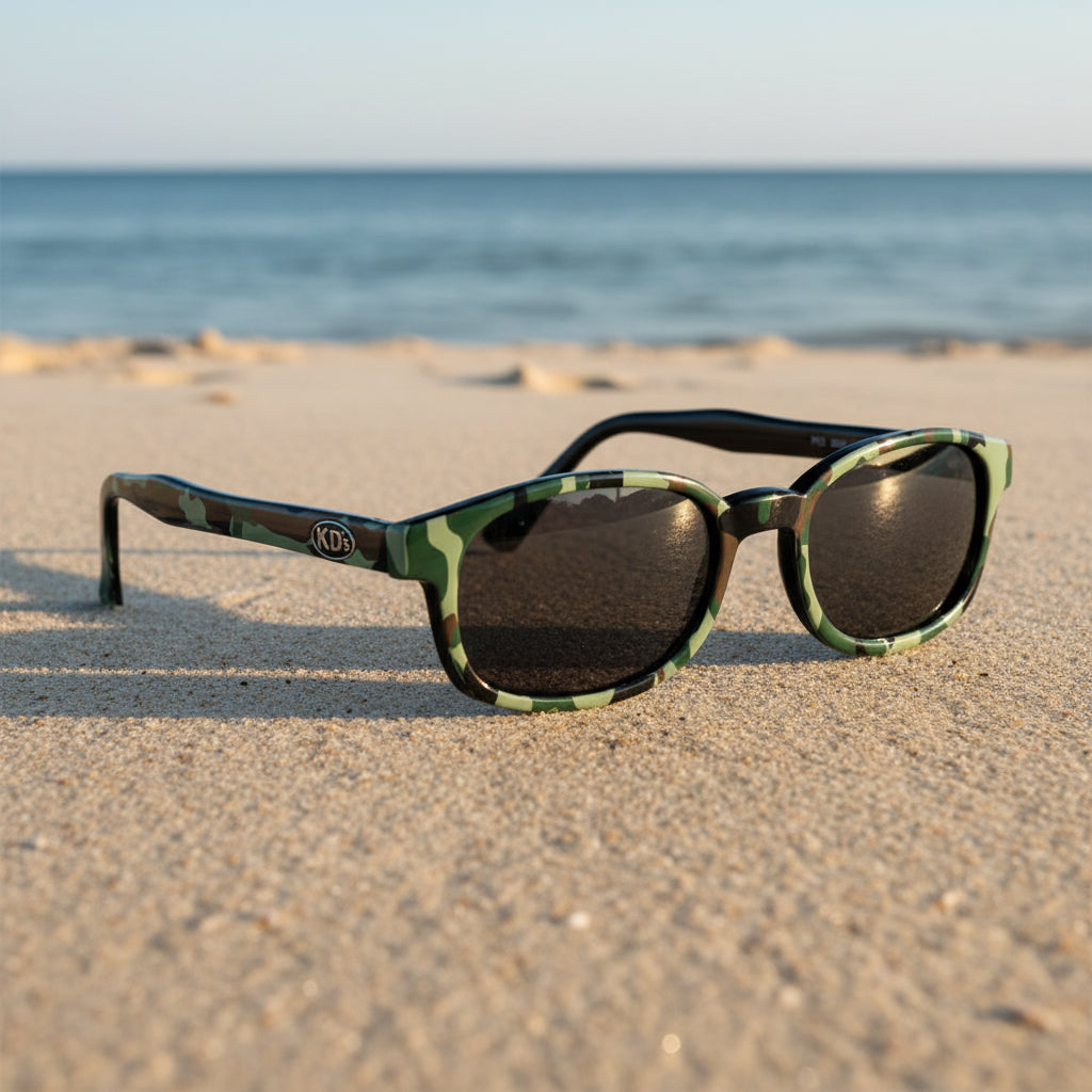 Lunettes de soleil KD's 2021 à monture camouflage militaire et verres fumés posées sur une plage de sable face à la mer.
