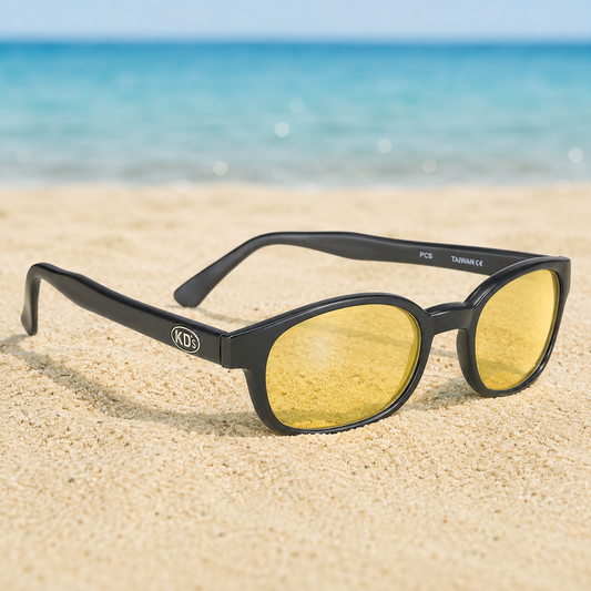 Lunettes de soleil KD's 21112 à monture noir mat et verres jaunes posées sur une plage de sable face à la mer.