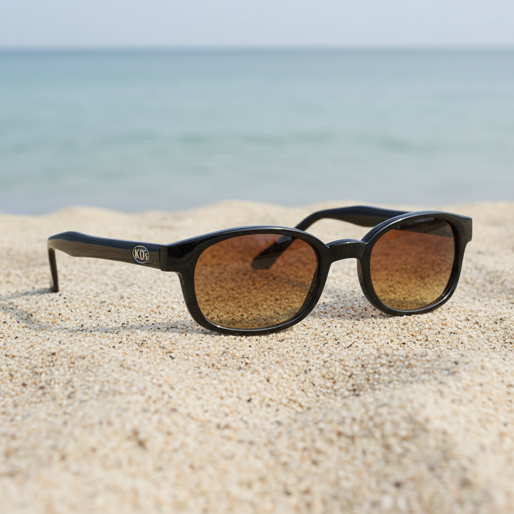 Lunettes de soleil KD's 21119 à monture noir brillant et verres ambre posées sur une plage de sable face à la mer.