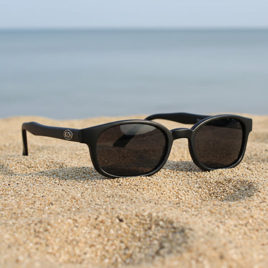 Lunettes de soleil KD's 21120 à monture noir mat et verres fumés posées sur une plage de sable face à la mer.