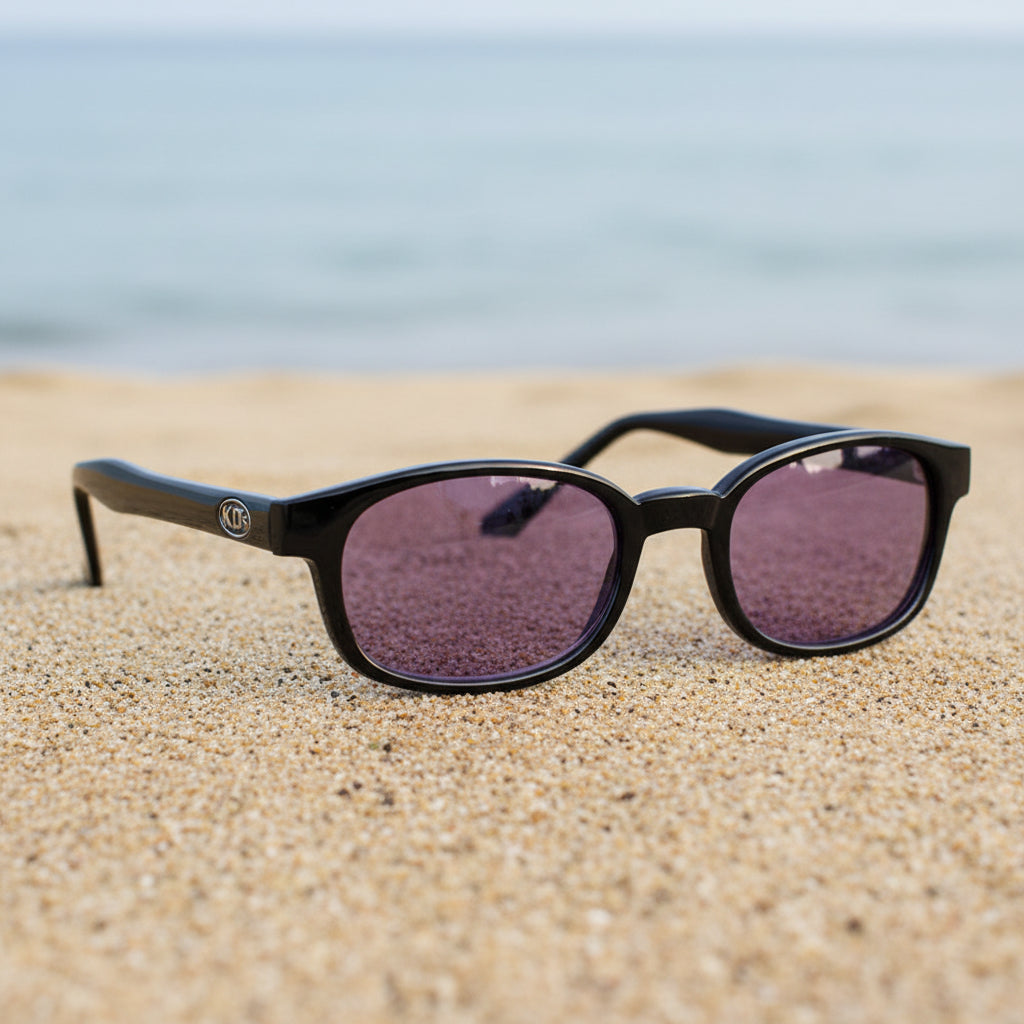 Lunettes de soleil KD's 21216 à monture noir brillant et verres violets posées sur une plage de sable face à la mer.