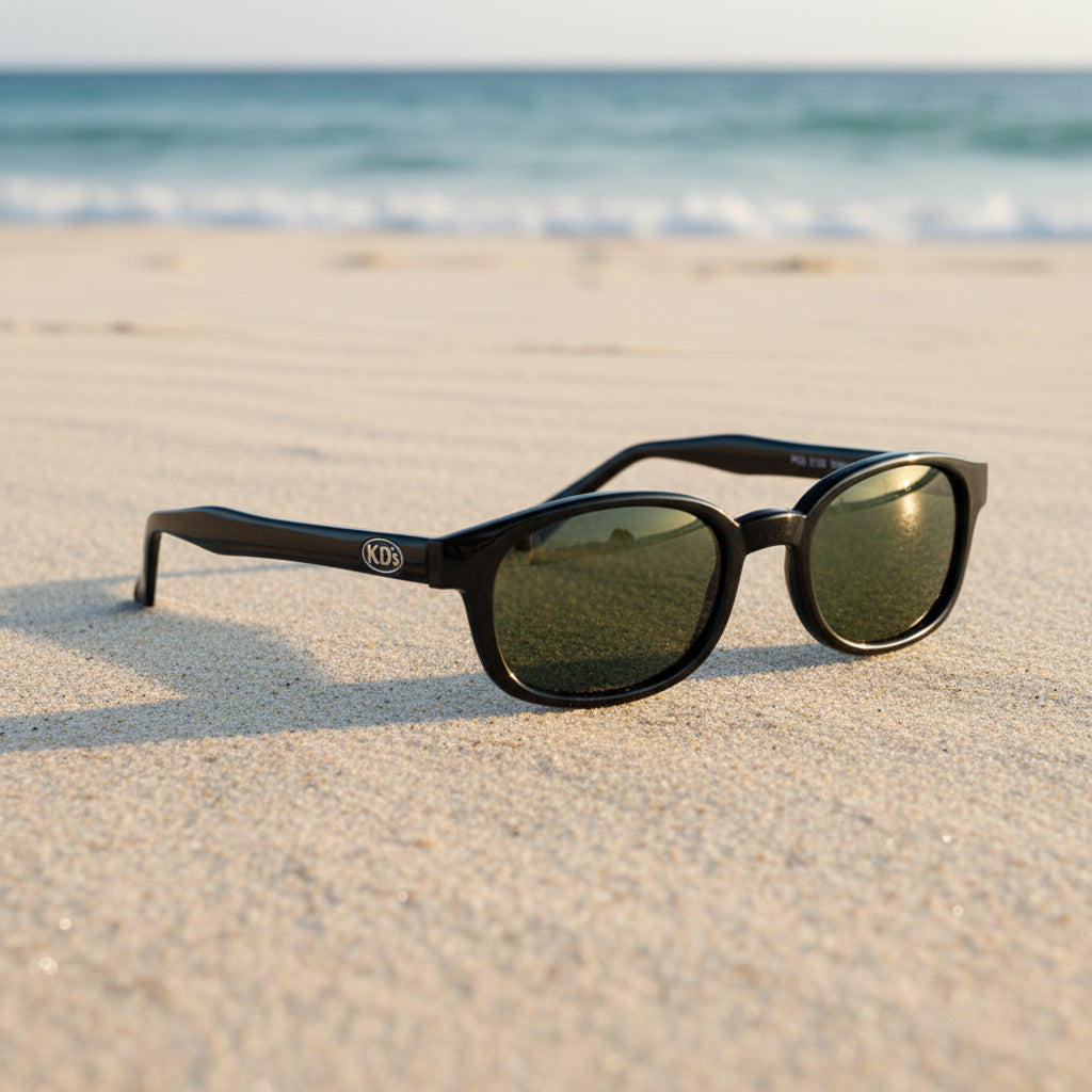 Lunettes de soleil KD's 2126 à monture noir brillant et verres vert foncé posées sur une plage de sable face à la mer.
