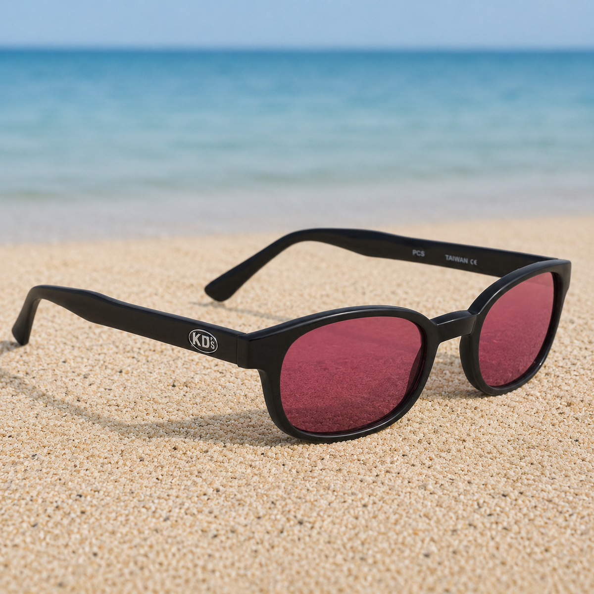 Lunettes de soleil KD's 22120 à monture noir mat et verres rose foncé posées sur une plage de sable face à la mer.