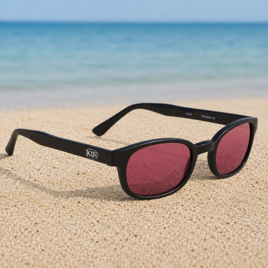 Lunettes de soleil KD's 22120 à monture noir mat et verres rose foncé posées sur une plage de sable face à la mer.