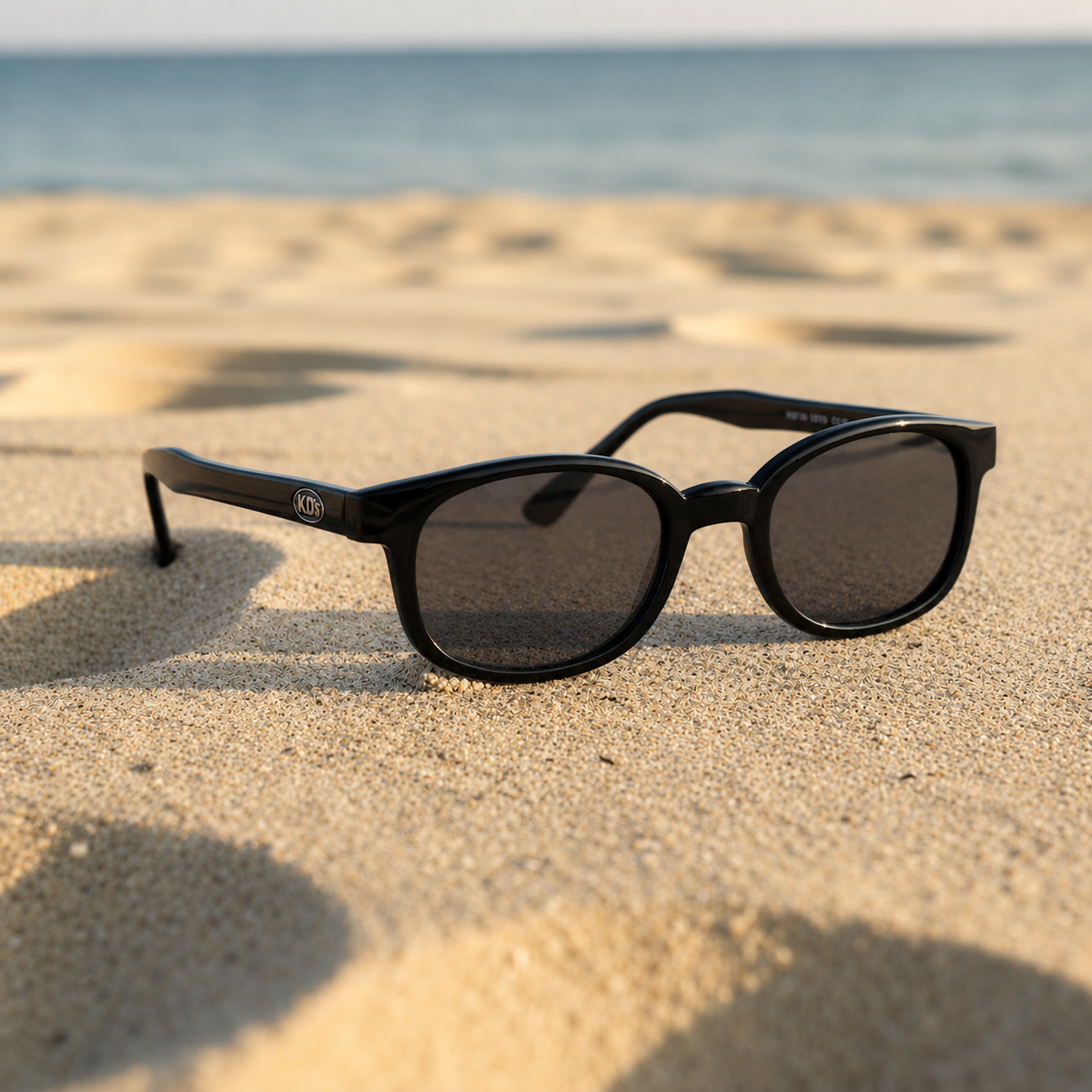 Lunettes de soleil X-KD's 10019 polarisées à monture noir brillant et verres fumés posées sur une plage de sable face à la mer.
