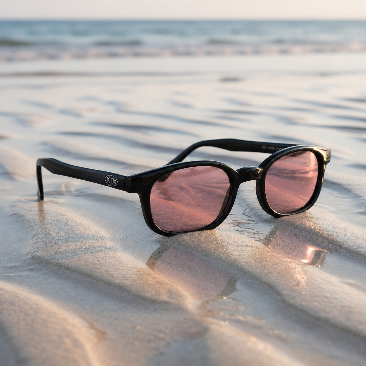 Lunettes de soleil X-KD's 10120 à monture noir brillant et verres roses posées sur une plage de sable face à la mer.