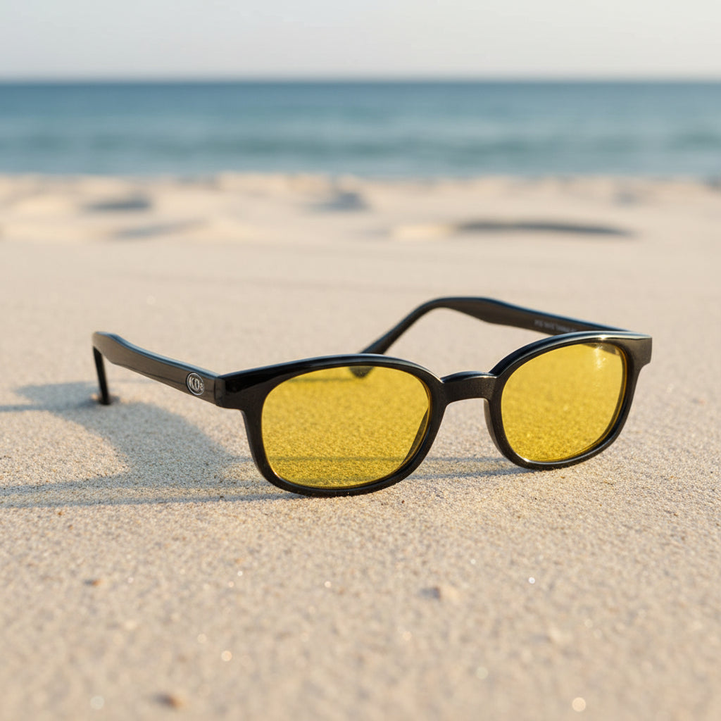 Lunettes de soleil X-KD's 10129 à monture noir brillant et verres jaunes polarisés posées sur une plage de sable face à la mer.