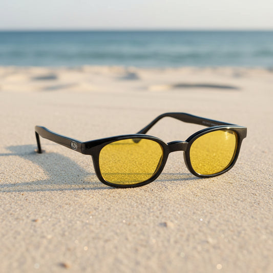 Lunettes de soleil X-KD's 10129 à monture noir brillant et verres jaunes polarisés posées sur une plage de sable face à la mer.