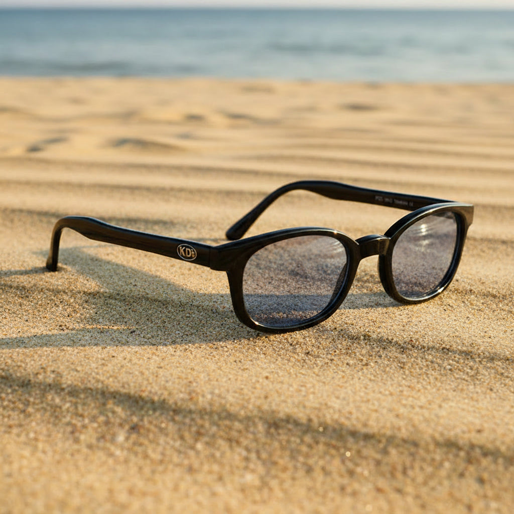 Lunettes de soleil X-KD's 1012 à monture noir brillant et verres bleus miroir posées sur une plage de sable face à la mer.