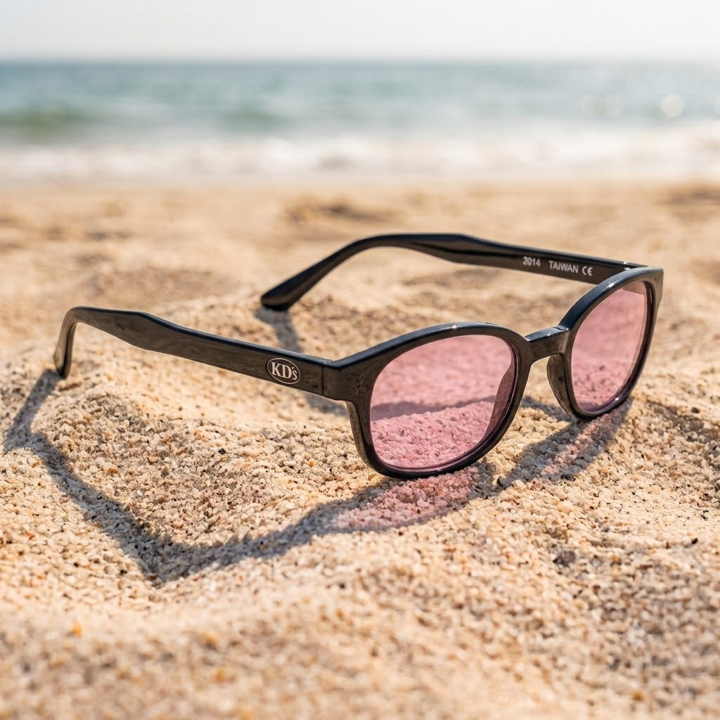 Lunettes X-KD's 1014 grande taille à monture noir brillant et verres rose clair posées sur une plage de sable face à la mer.