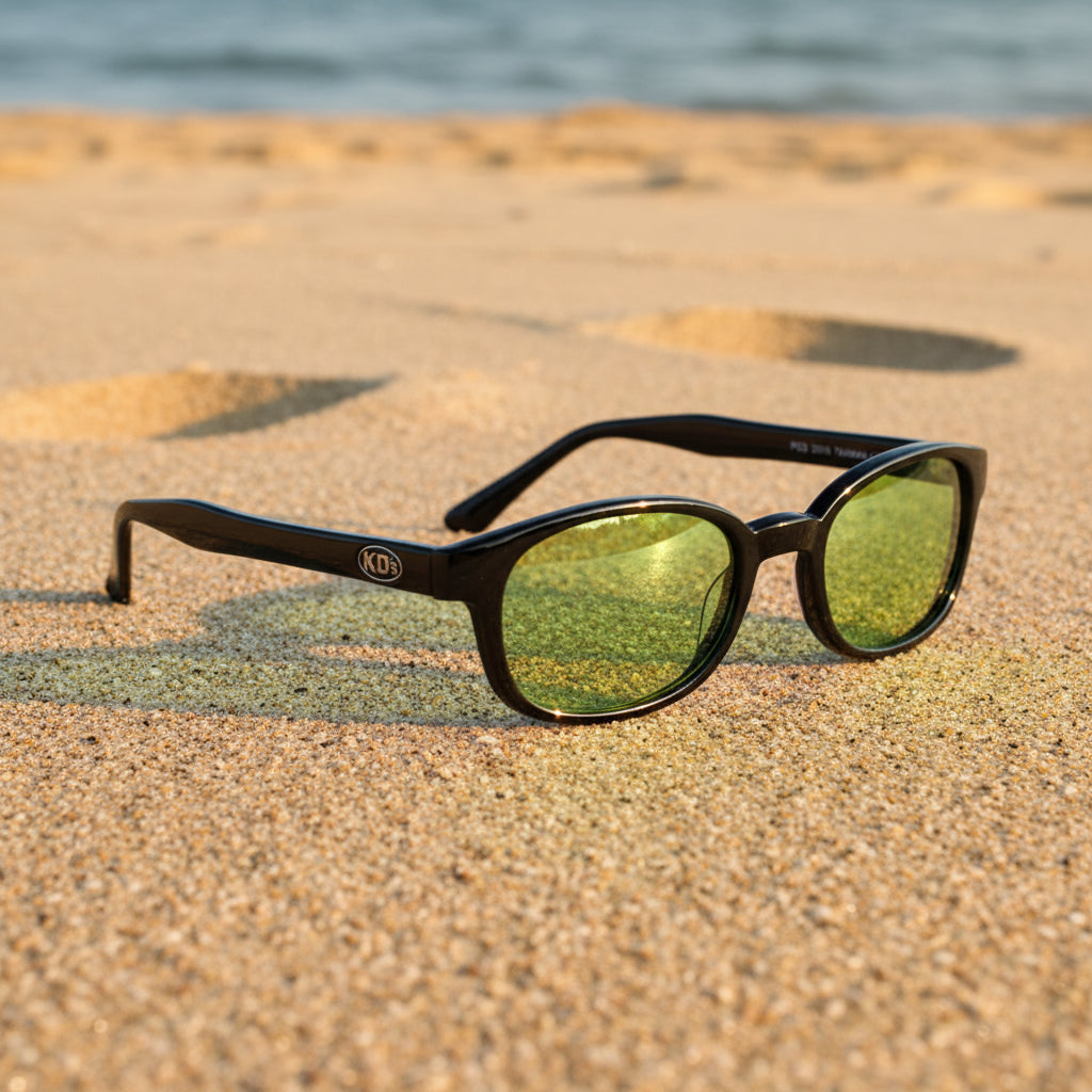 Lunettes de soleil X-KD's 1016 à monture noir brillant et verres verts posées sur une plage de sable face à la mer.