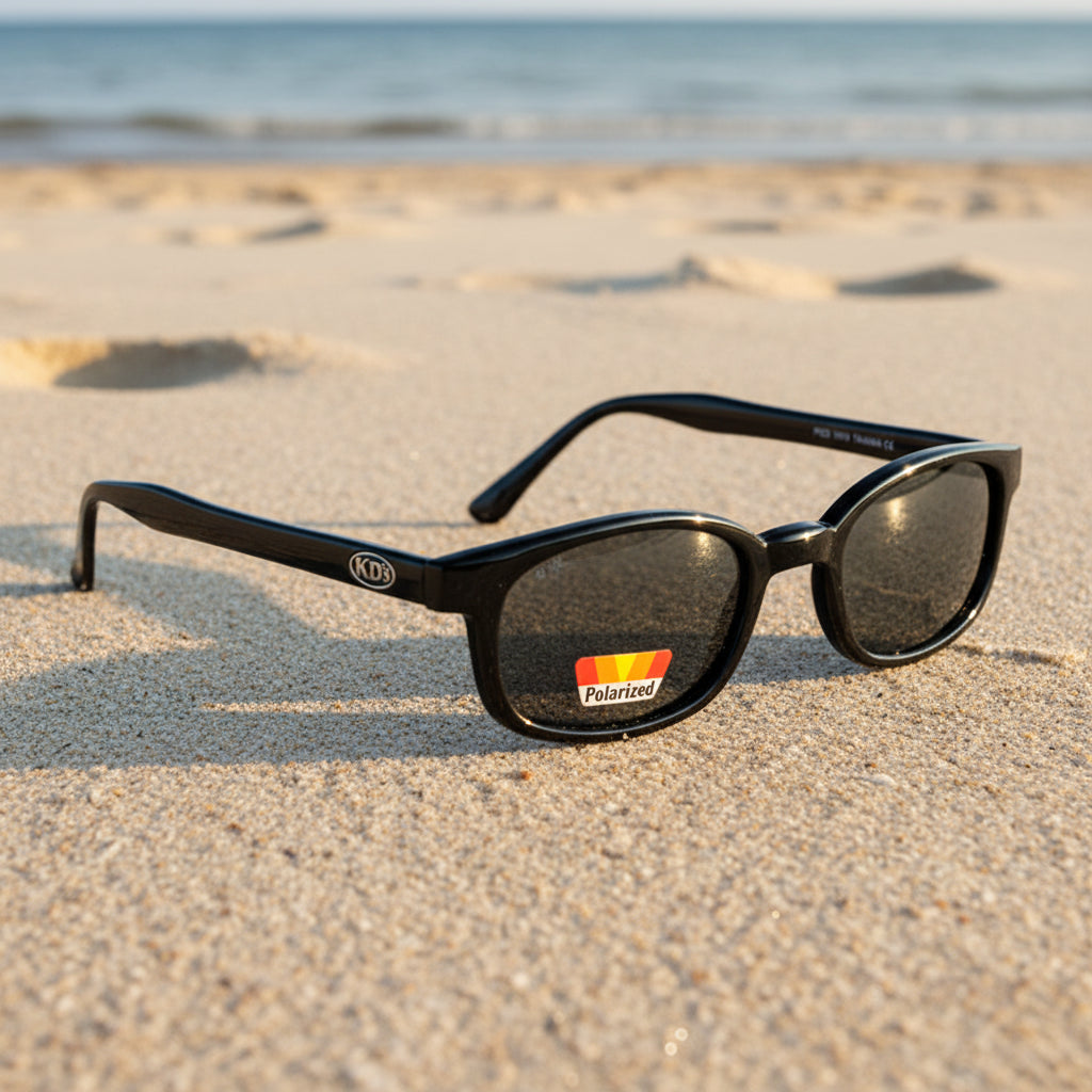 Lunettes de soleil X-KD's 1019 à monture noir brillant et verres gris polarisés posées sur une plage de sable face à la mer.