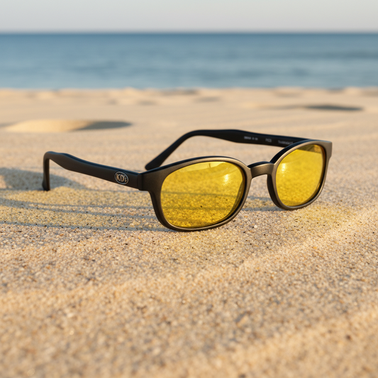 Lunettes de soleil X-KD's 11112 à monture noir mat et verres jaunes posées sur une plage de sable face à la mer.