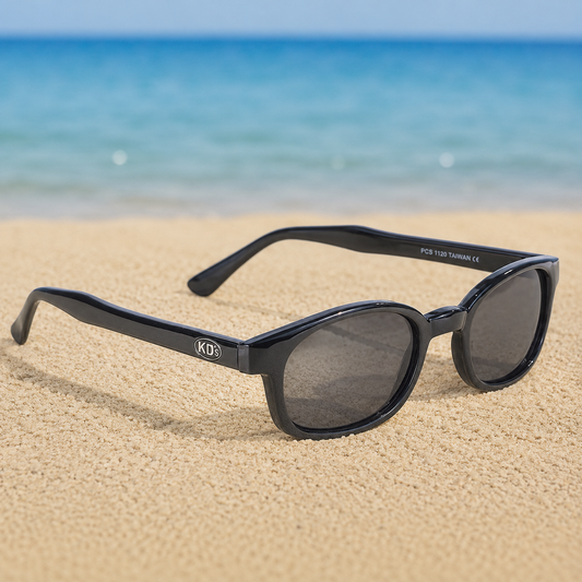 Lunettes de soleil X-KD's 1120 grande taille à monture noir brillant et verres gris foncé posées sur une plage de sable face à la mer.
