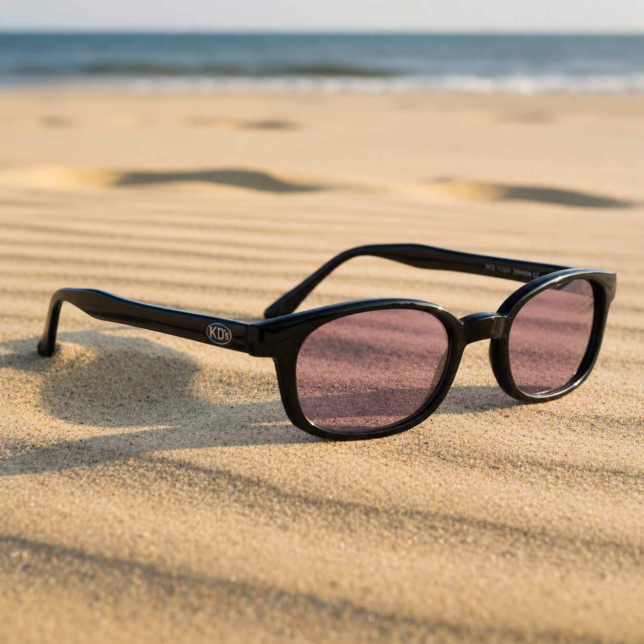 Lunettes de soleil X-KD's 11216 à monture noir brillant et verres violets posées sur une plage de sable face à la mer.