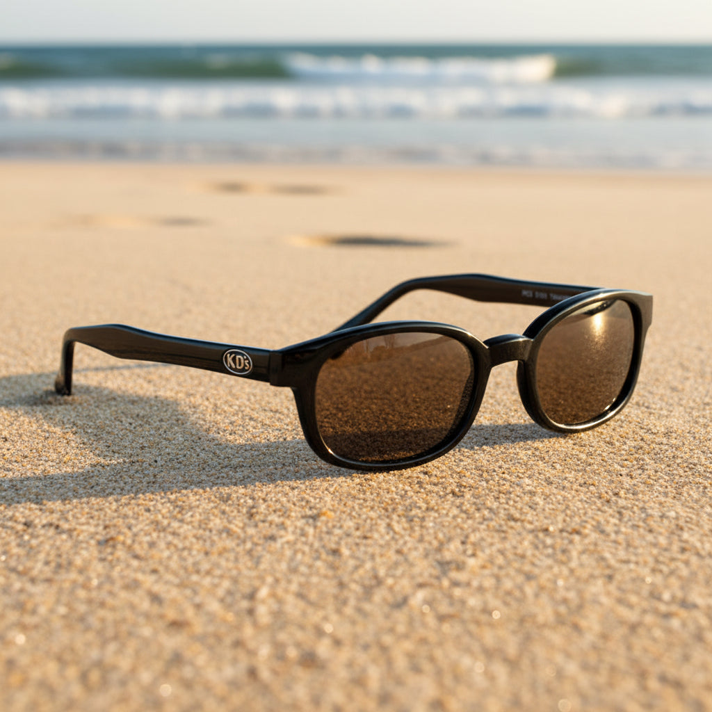 Lunettes de soleil X-KD's 1121 à monture noir brillant et verres marron posées sur une plage de sable face à la mer.