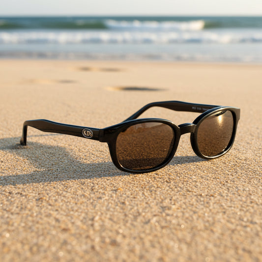 Lunettes de soleil X-KD's 1121 à monture noir brillant et verres marron posées sur une plage de sable face à la mer.