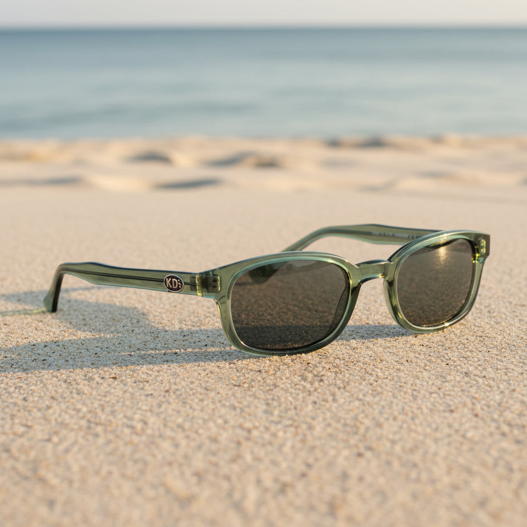 Lunettes de soleil X-KD's 11269 à monture vert translucide et verres verts polarisés posées sur une plage de sable face à la mer.