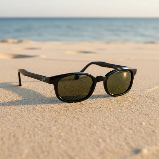 Lunettes de soleil X-KD's 1126 à monture noir brillant et verres vert foncé posées sur une plage de sable face à la mer.