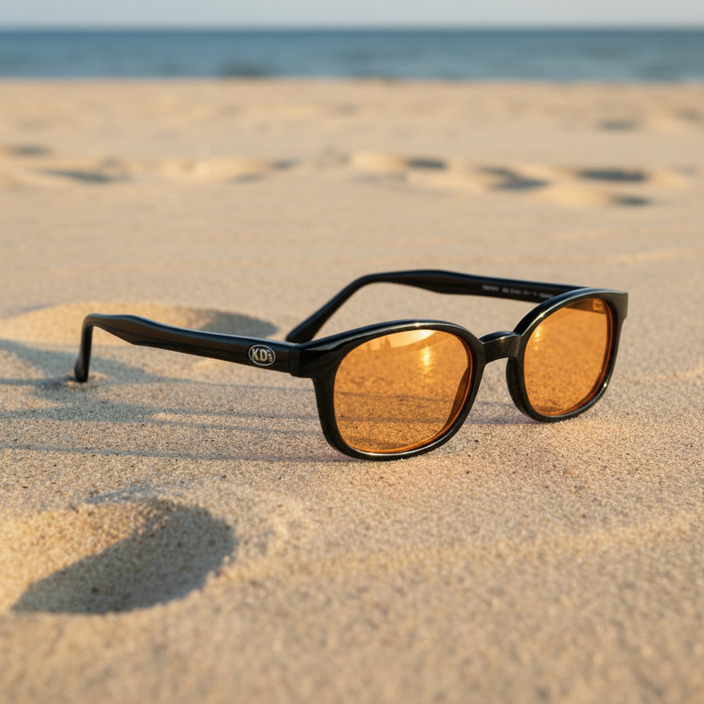 Lunettes de soleil X-KD's 1128 à monture noir brillant et verres orange posées sur une plage de sable face à la mer.
