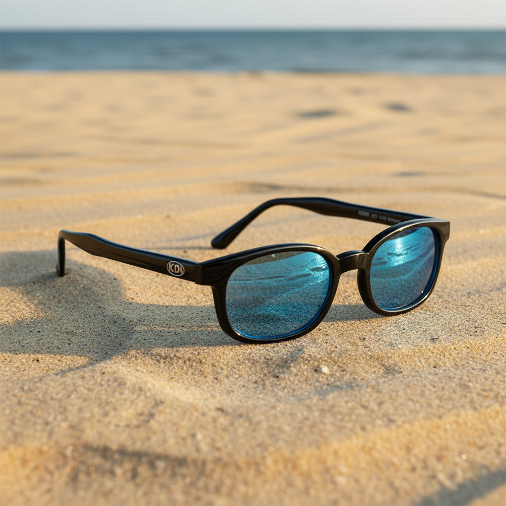 Lunettes de soleil X-KD's 1129 à monture noir brillant et verres bleu turquoise posées sur une plage de sable face à la mer.