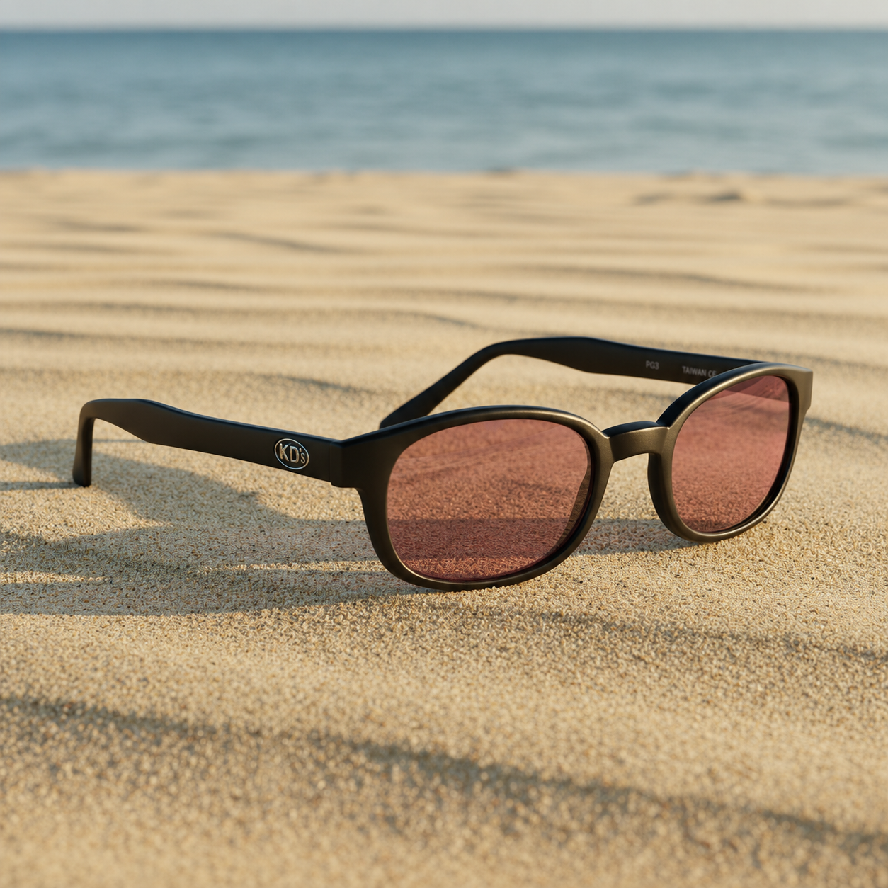 Lunettes de soleil X-KD's 12120 à monture noir mat et verres rose foncé posées sur une plage de sable face à la mer.