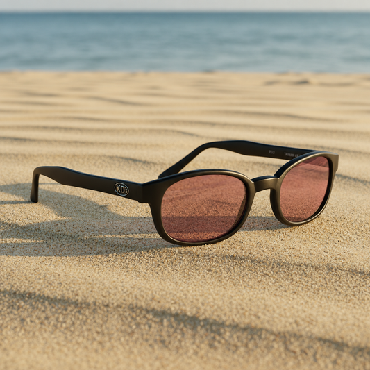 Lunettes de soleil X-KD's 12120 à monture noir mat et verres rose foncé posées sur une plage de sable face à la mer.