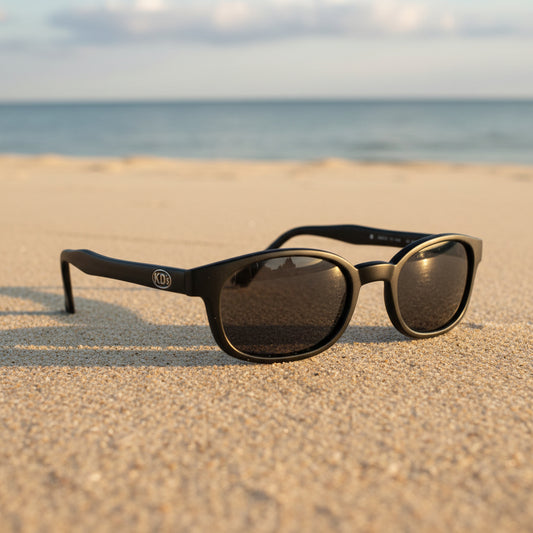 Lunettes de soleil X-KD's 20019 à monture noir mat et verres gris polarisés posées sur une plage de sable face à la mer.