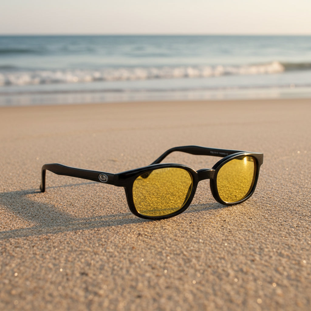 Lunettes de soleil X-KD's 10112 à monture noir brillant et verres jaunes posées sur une plage de sable face à la mer.