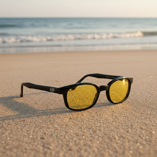 Lunettes de soleil X-KD's 10112 à monture noir brillant et verres jaunes posées sur une plage de sable face à la mer.