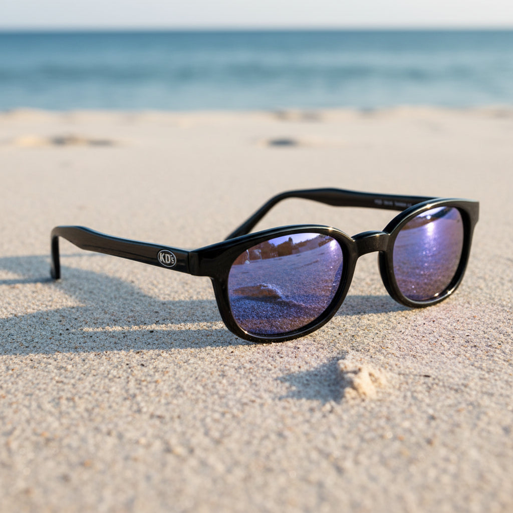 Lunettes de soleil X-KD's 10118 à monture noir brillant et verres miroir irisés posées sur une plage de sable face à la mer.