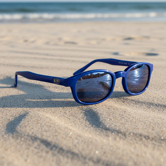 Lunettes de soleil X-KD's 10122 à monture bleu glacé et verres bleu miroir posées sur une plage de sable face à la mer.