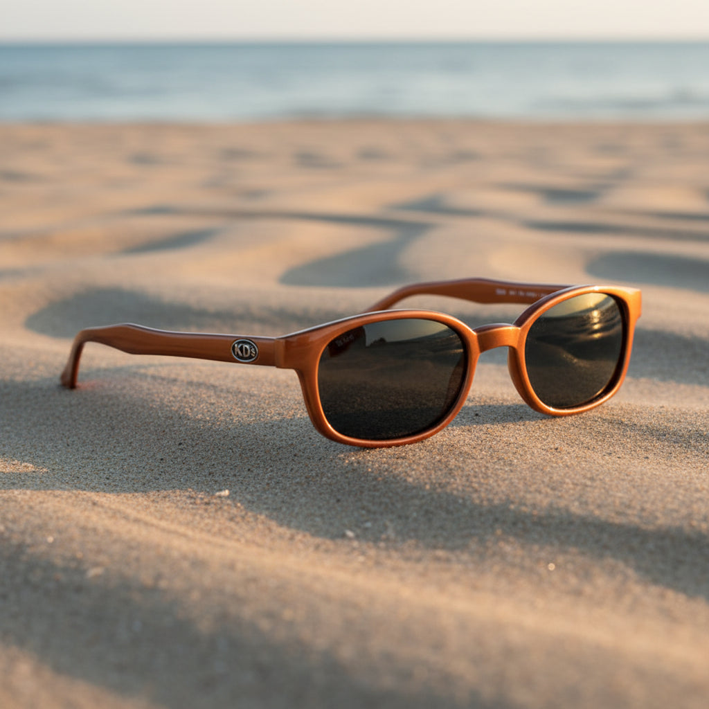 Lunettes de soleil X-KD's 10128 à monture orange métallisé et verres gris fumé posées sur une plage de sable face à la mer.