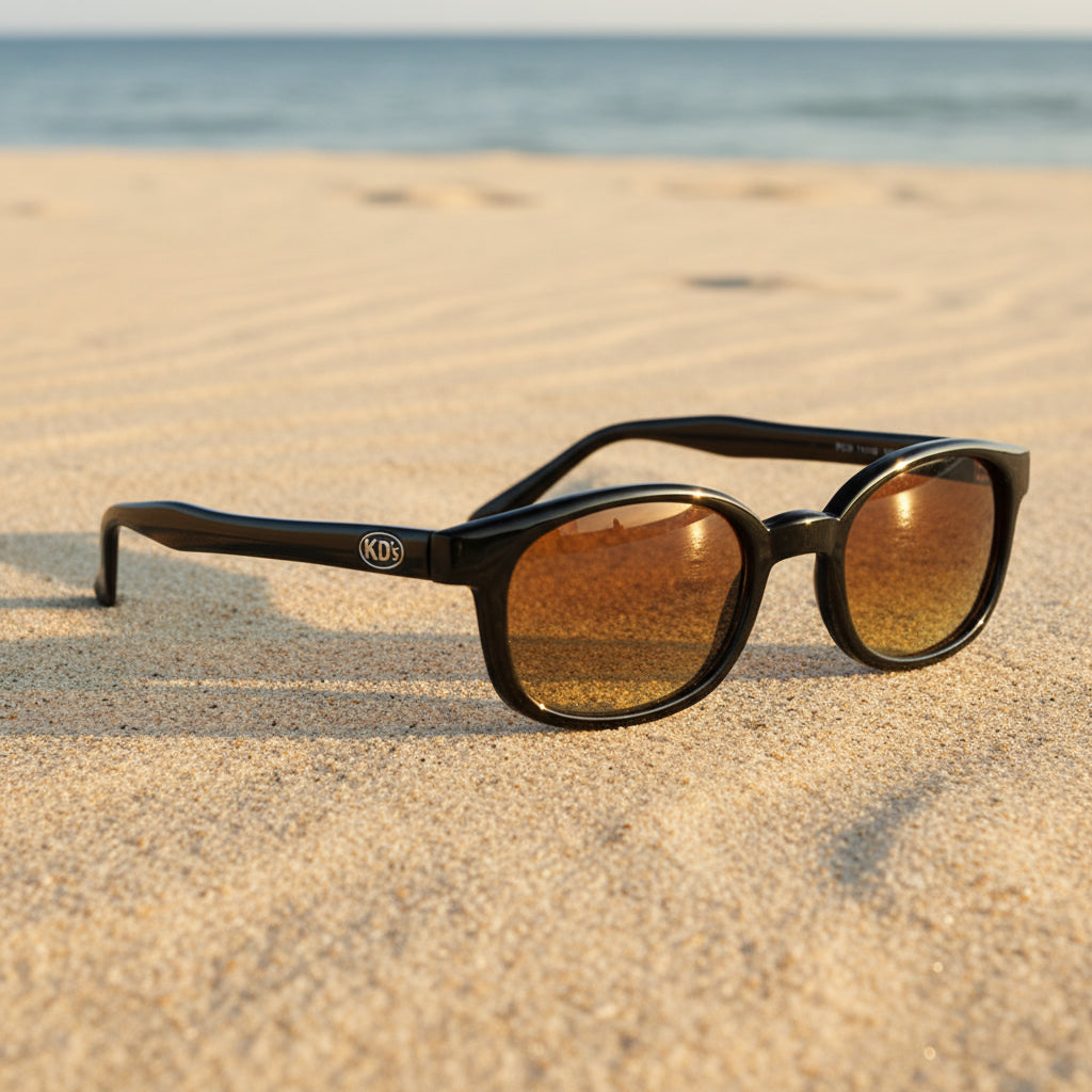 Lunettes de soleil X-KD's 11119 à monture noir brillant et verres ambre posées sur une plage de sable face à la mer.